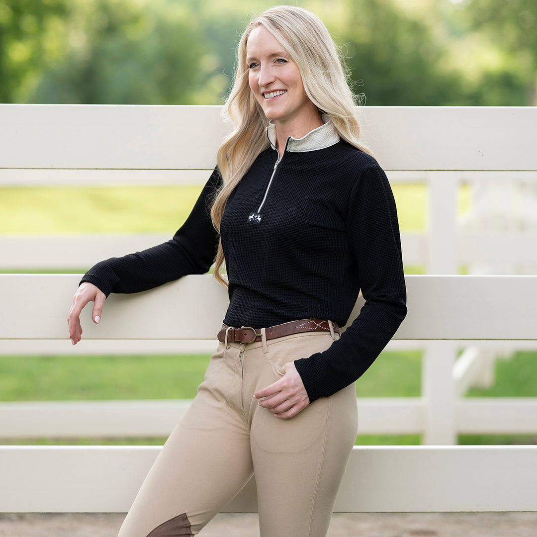 Woman in equestrian attire sitting on a white fence with greenery in the background