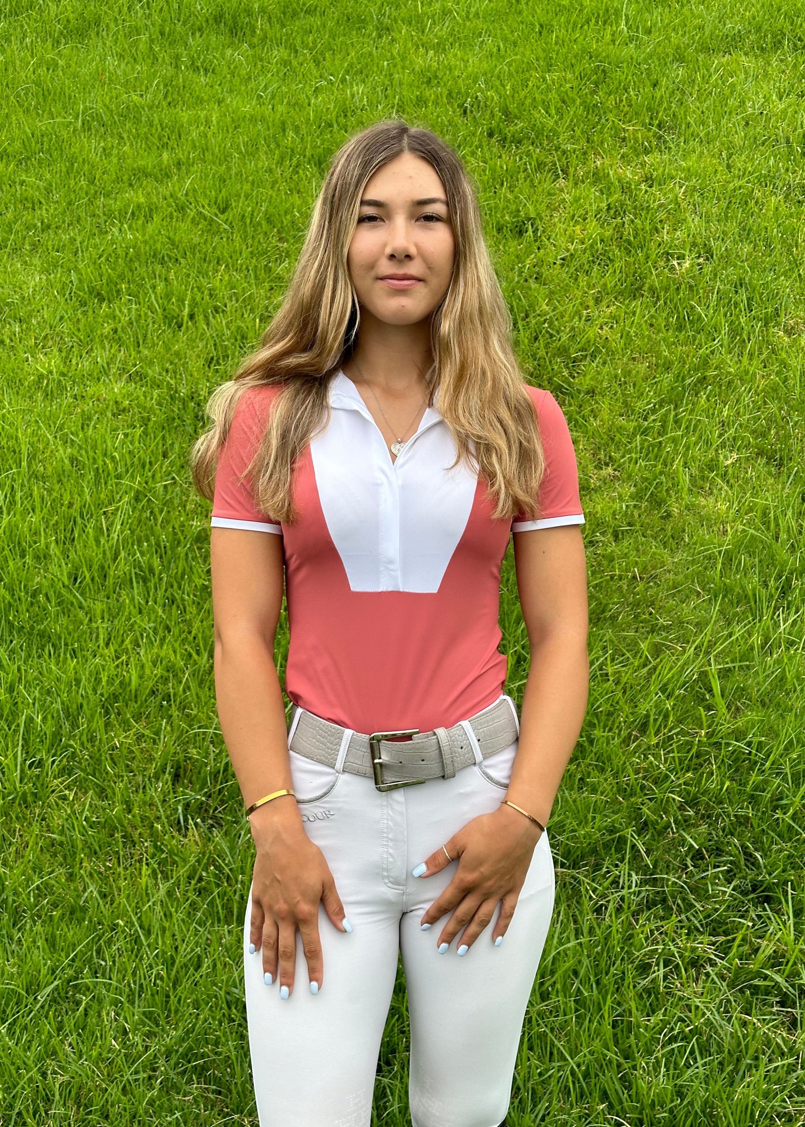 Woman wearing a pink equestrian shirt with a white collar and white pants standing on grass.