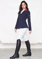 Woman wearing a navy equestrian jacket with red collar, white shirt, white pants, and black riding boots on a white background.