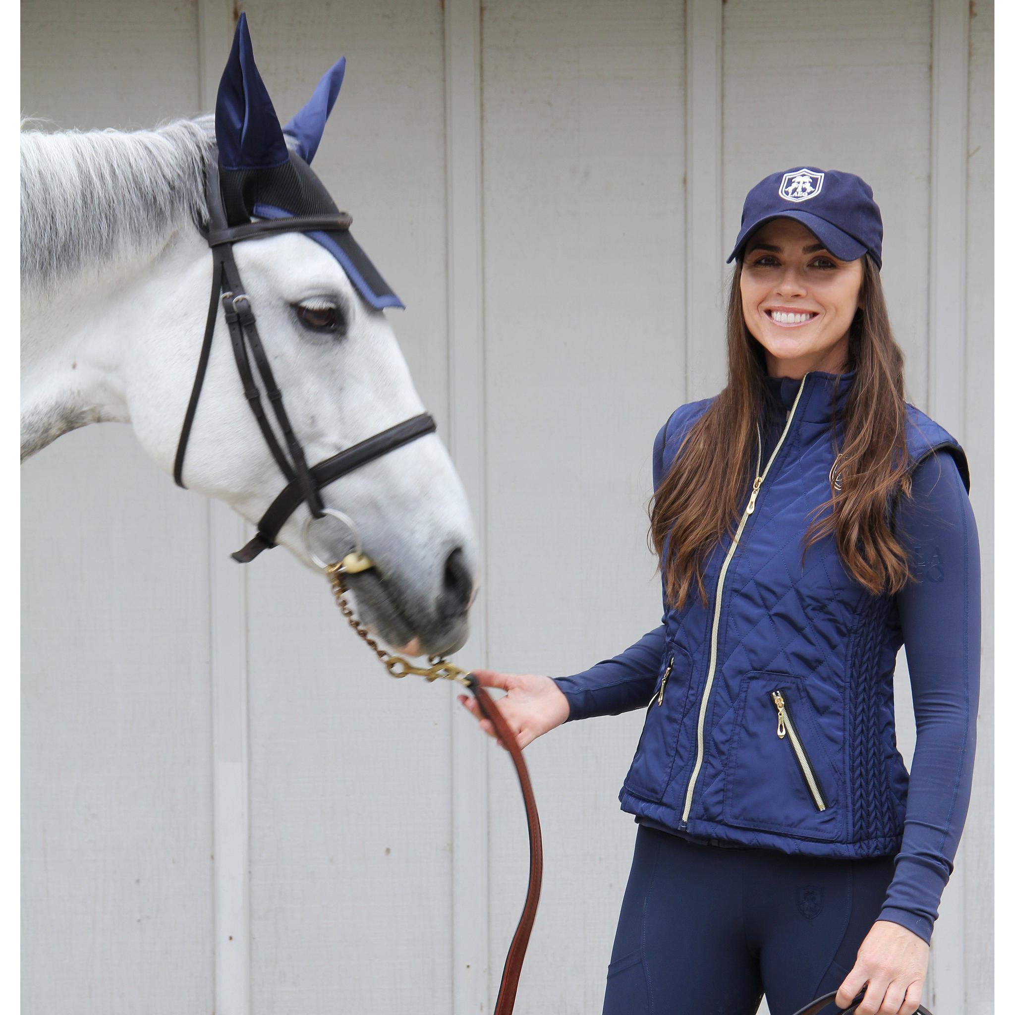 Woman in equestrian attire standing next to a white horse wearing a blue bridle.