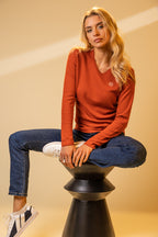 Woman sitting on a black stool wearing an orange sweater and blue jeans against a beige background