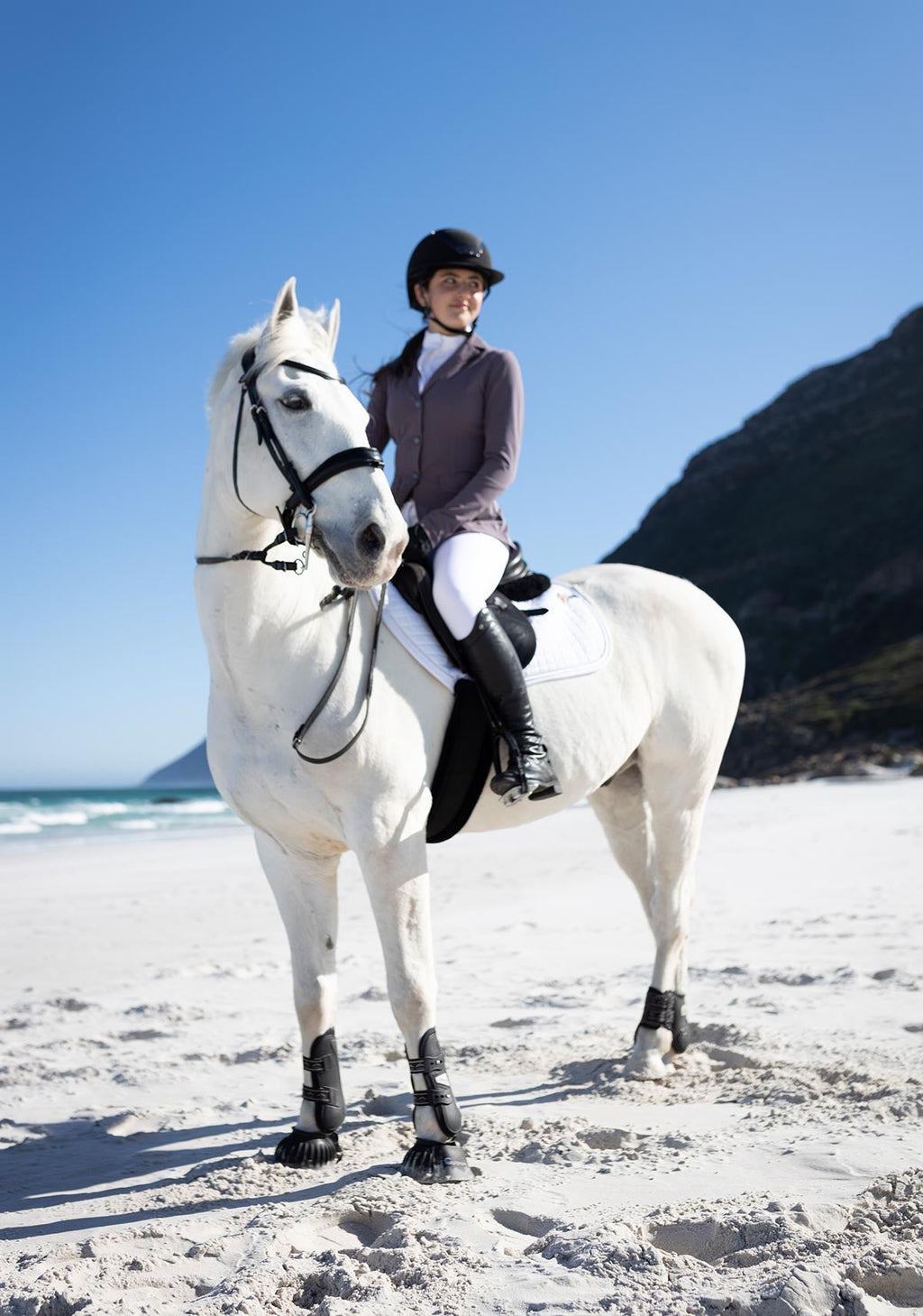 Person riding a white horse on a sandy beach with mountains in the background
