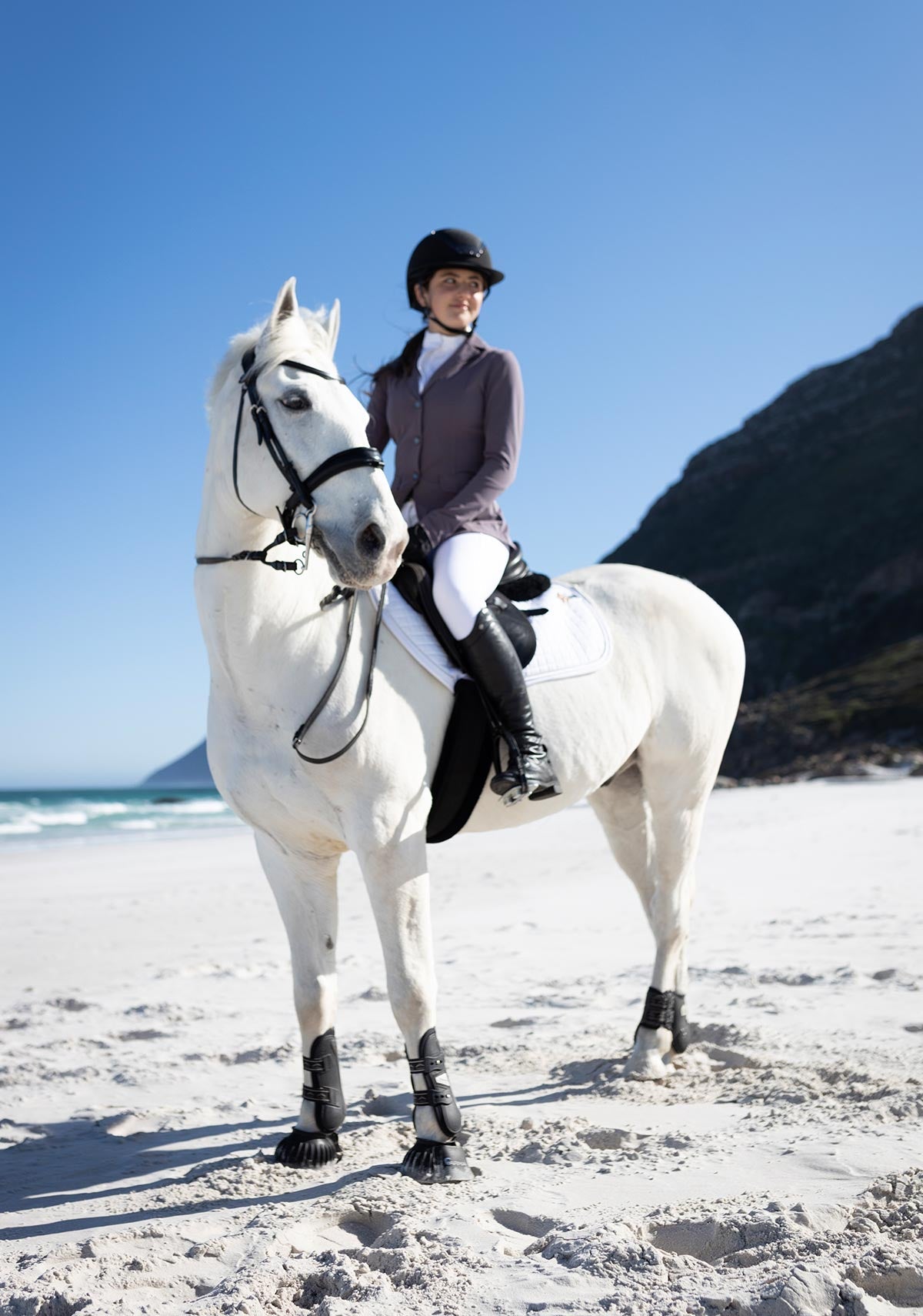 Person riding a white horse on a sandy beach with mountains in the background