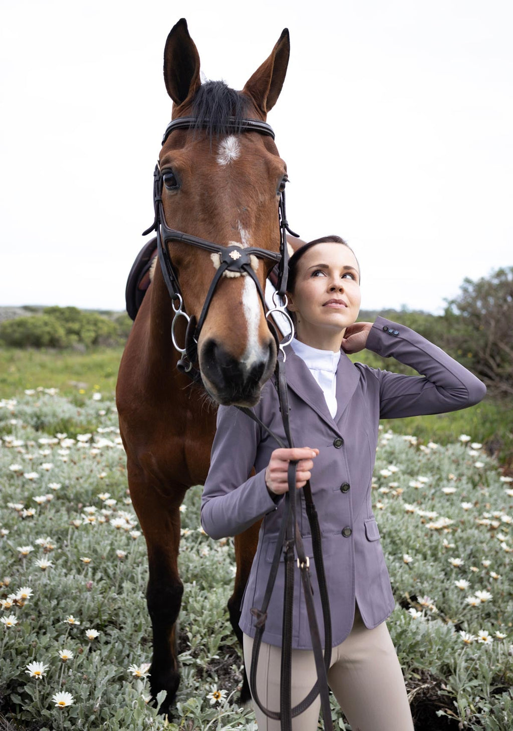 Woman in equestrian attire standing next to a horse in a field with flowers