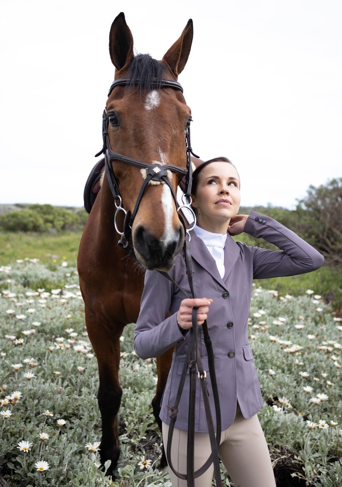 Woman in equestrian attire standing next to a horse in a field with flowers