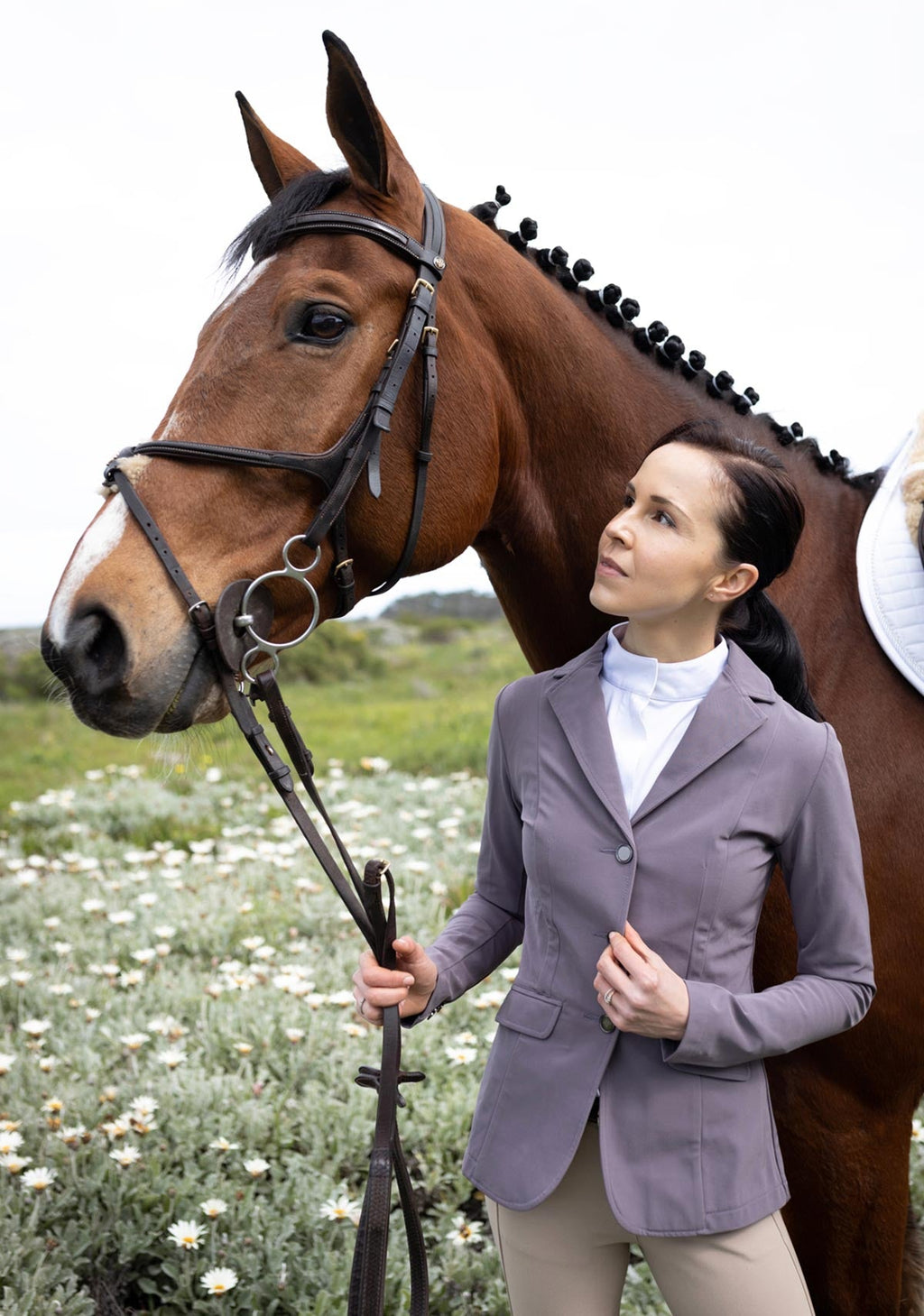 Woman in equestrian attire standing next to a horse in a field with flowers.