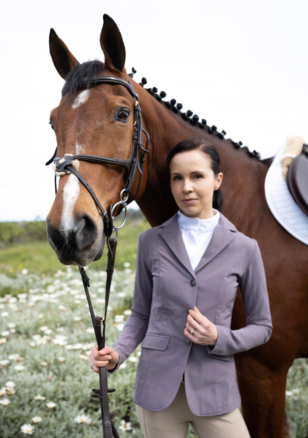 Woman in equestrian attire standing next to a brown horse in an outdoor setting.