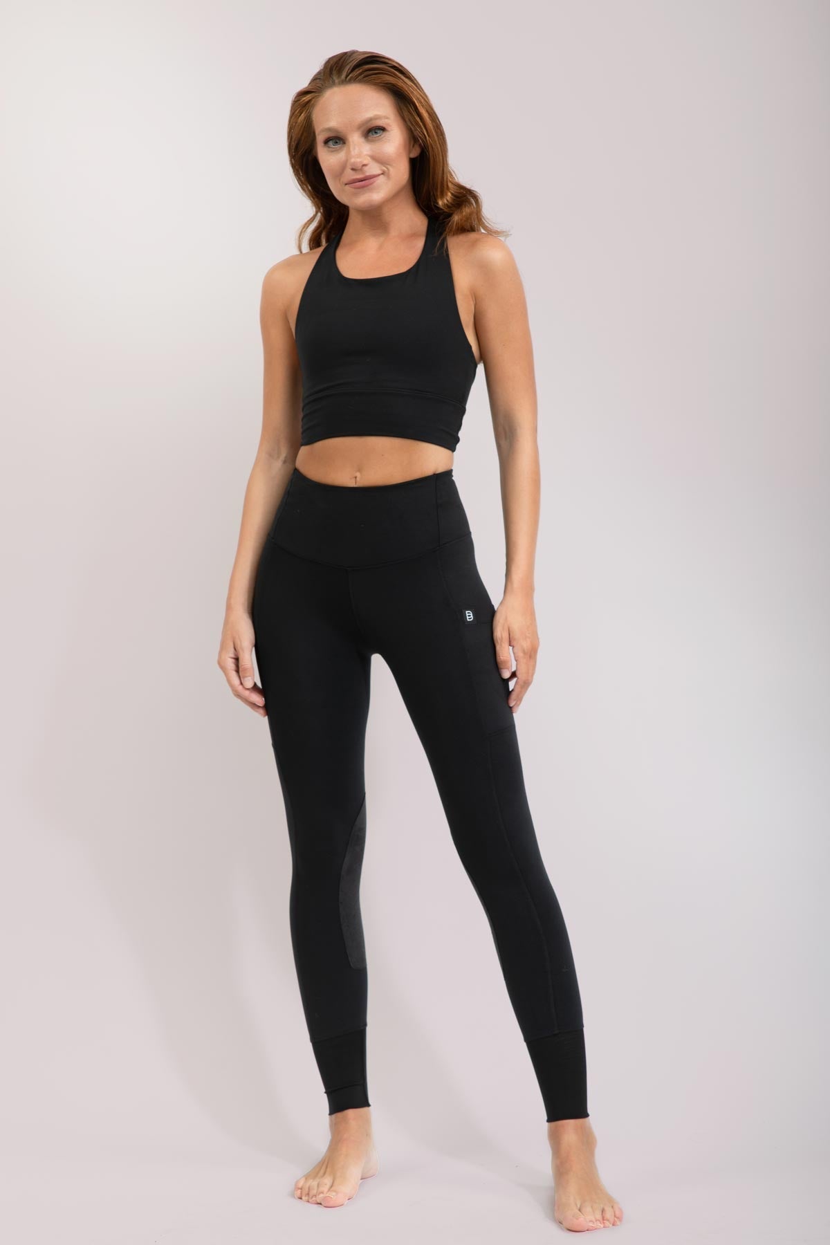 Woman wearing black equestrian riding leggings and sports bra on a light gray background