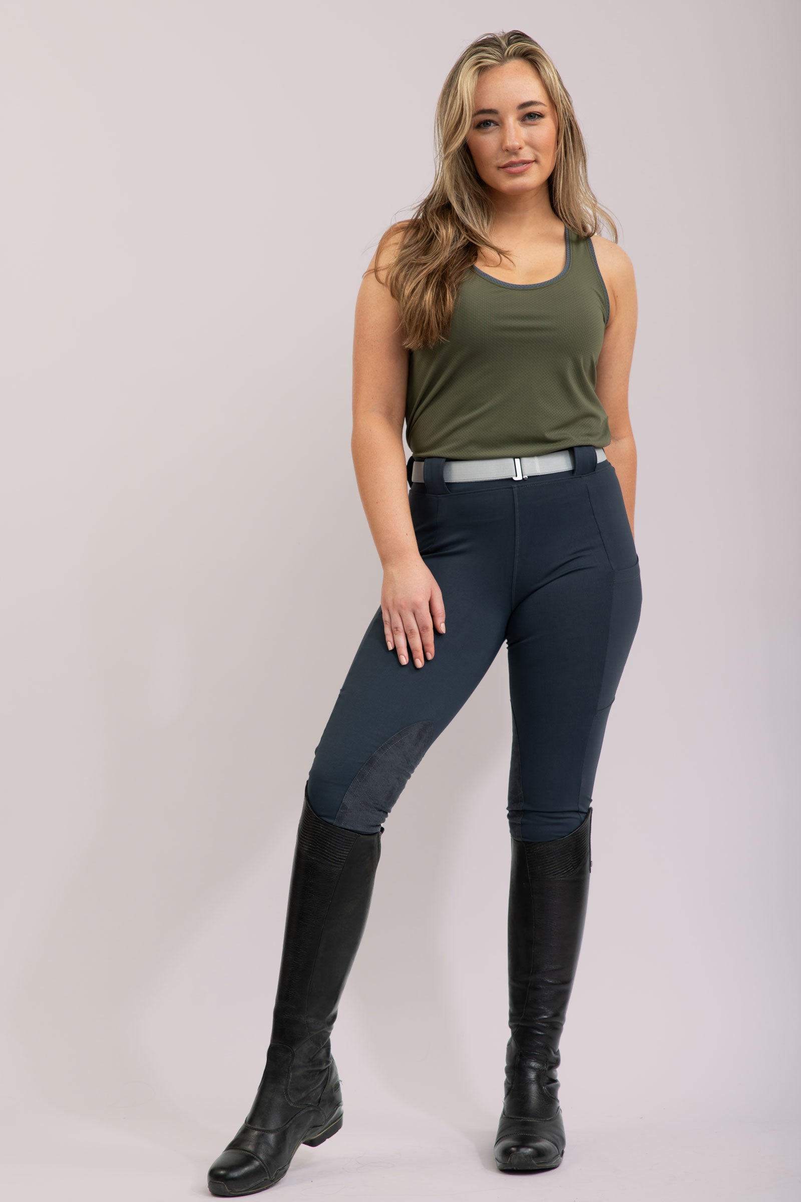 Woman wearing green tank top and dark riding pants with black boots on a plain background