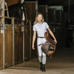 Woman in equestrian attire holding a saddle in a stable.