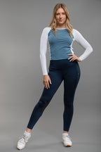 Woman wearing a blue top and dark leggings with a white long-sleeve shirt underneath on a gray background