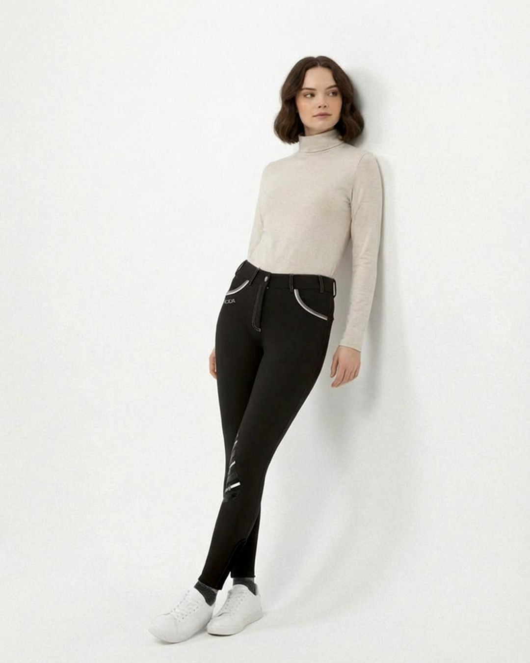 Woman wearing a beige sweater and black breeches against a white background