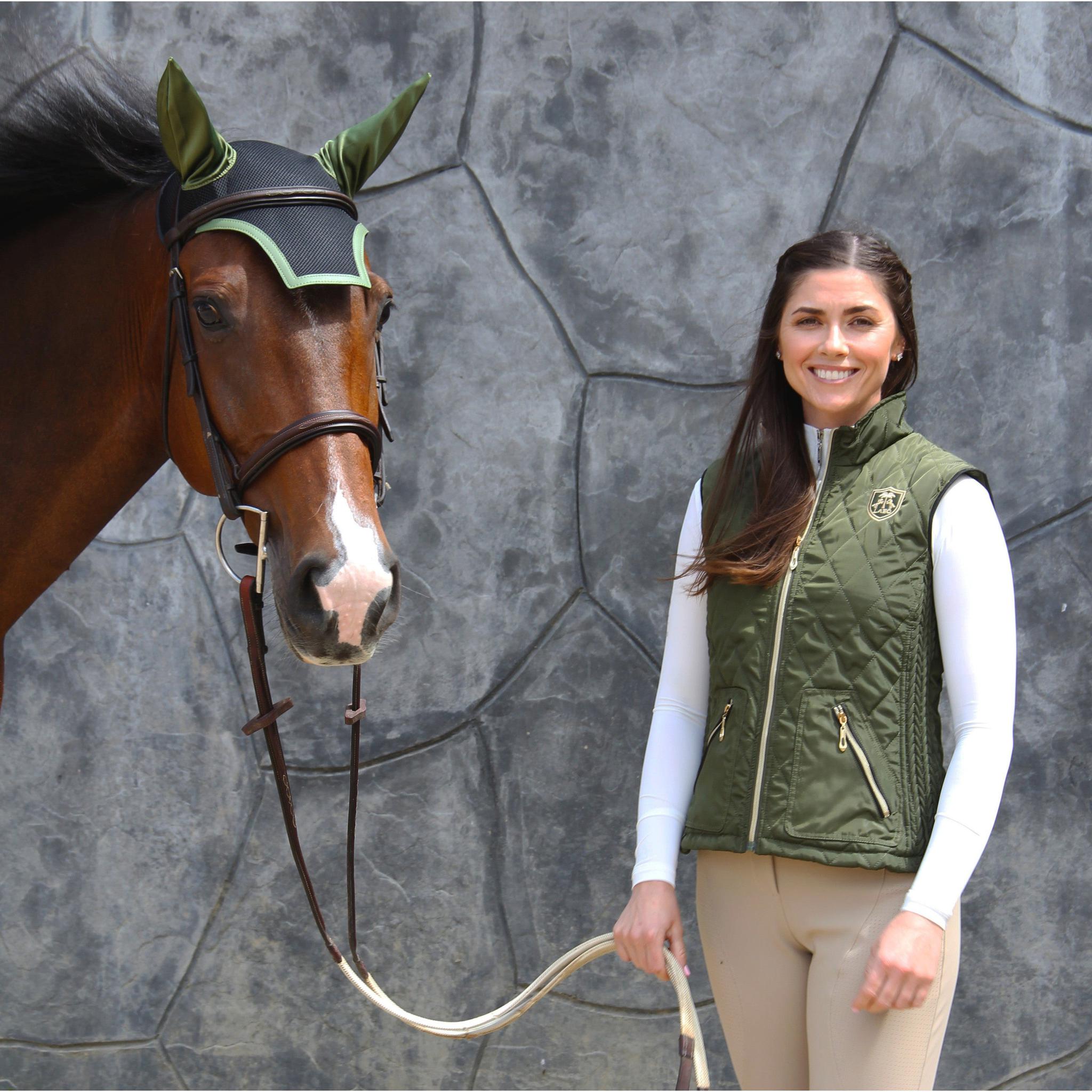 Woman in equestrian attire standing next to a horse with a bridle against a stone wall background