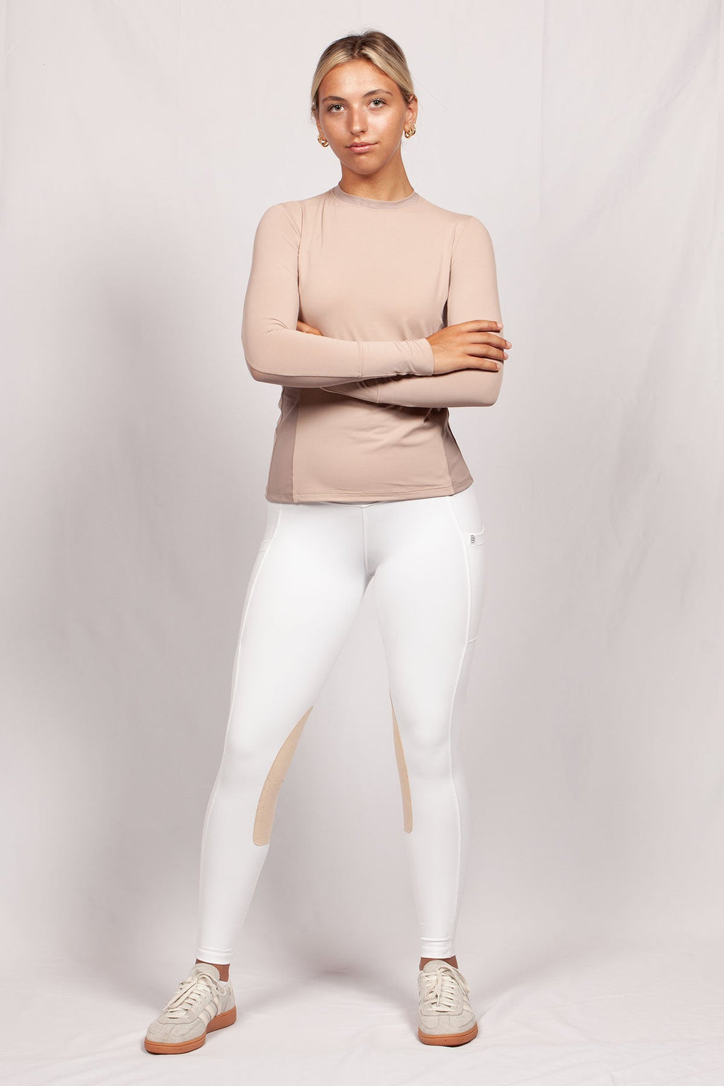Woman wearing a beige long-sleeve top and white leggings on a light gray background